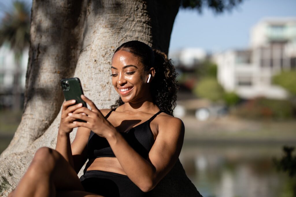 Happy woman in activewear using her phone outdoors by a tree.