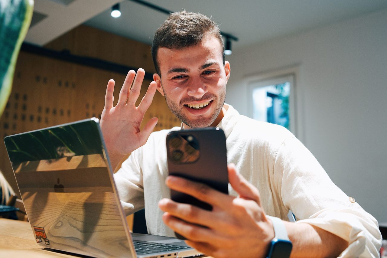 Smiling young man making a video call on his smartphone while sitting with a laptop in a modern office.